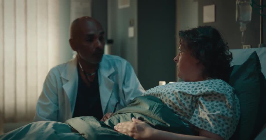 A male doctor checks in on a female patient in a hospital looking at the chart her feet and talking with her shaking his head no before getting up