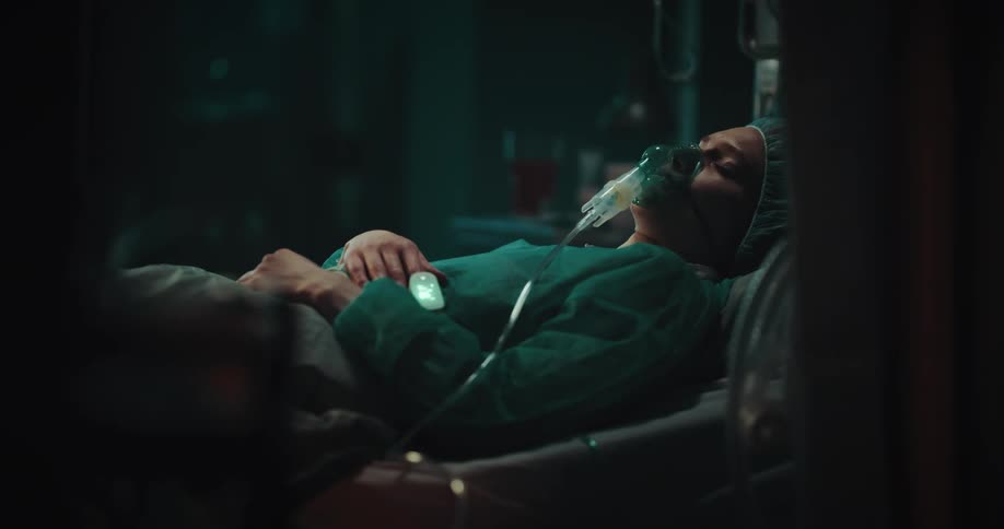 Woman in hospital gown lays in bed with tubes and oxygen mask