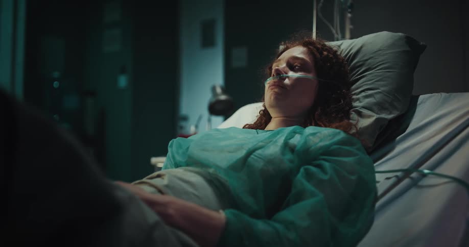 A woman with an oxygen mask on a verge of tears as she reclines in a hospital bed