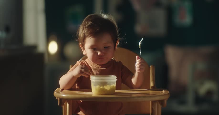 The little child is in the highchair and they are holding a spoon but eating from the bowl in front of them with their empty hand