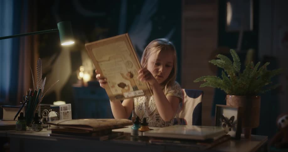 A young blonde girl sits at a desk and opens a book to read