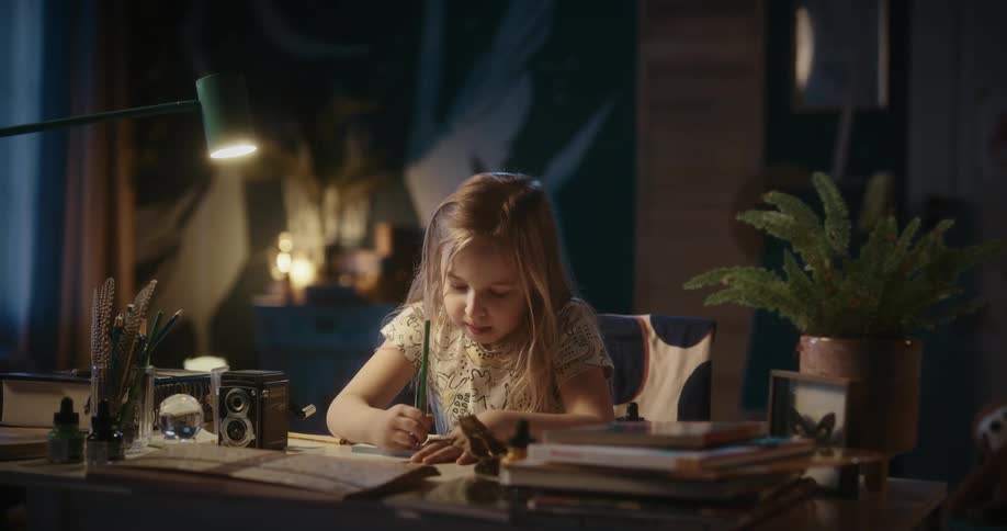 A young blonde girl sits at a desk and writes on a tablet of paper
