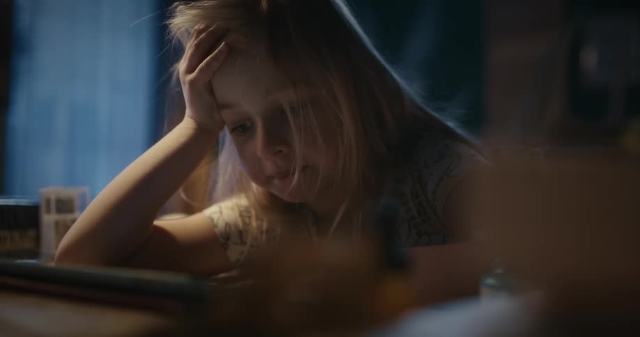 A little blonde girl is watching something on the screen of electronic equipment on a table