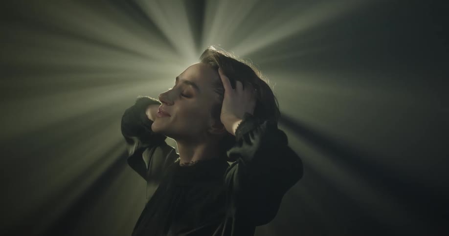 A young female model is standing and blocking the light in a dark space as she touches her head with her hands
