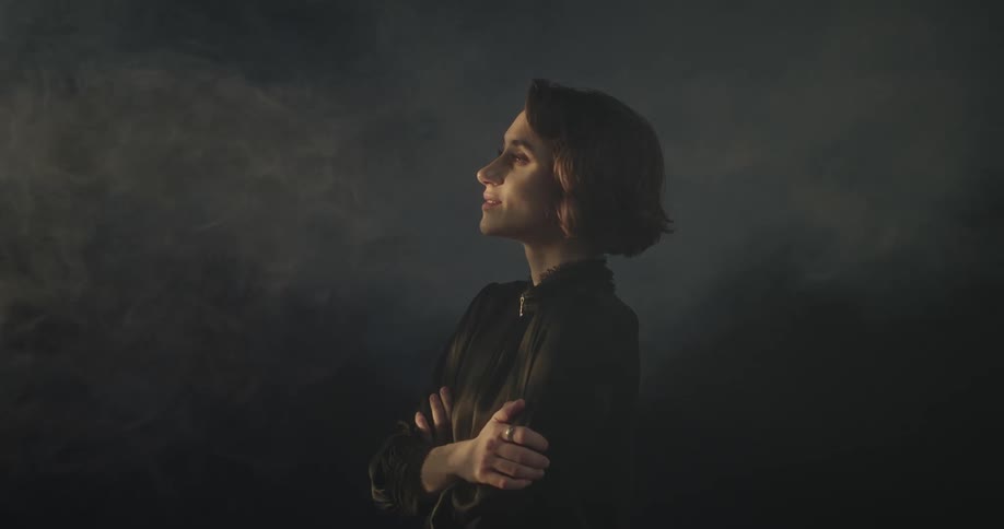 A woman is standing with her arms crossed and smiling while a light illuminates the dark and smoky environment