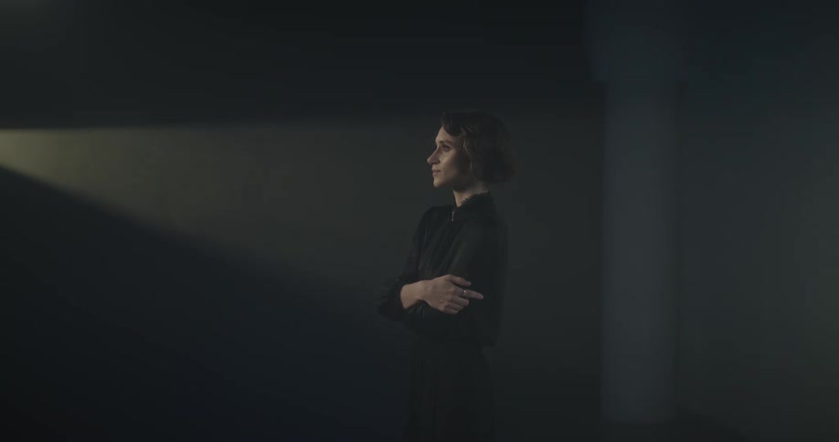 A woman with arms crossed stands facing a projection light that slowly moves projecting a moving square box of light on the wall behind her