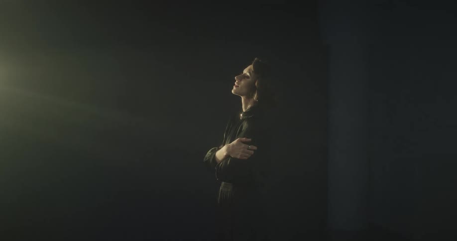 A female model standing in a dark room wearing all black as light shines and move in the room