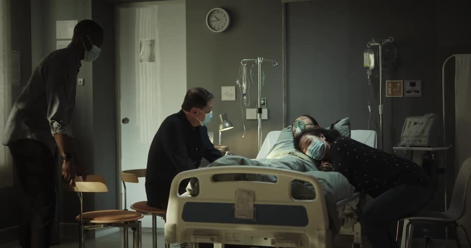 Family around a woman sleeping in a hospital bed in a hospital room and they are caressing the patient