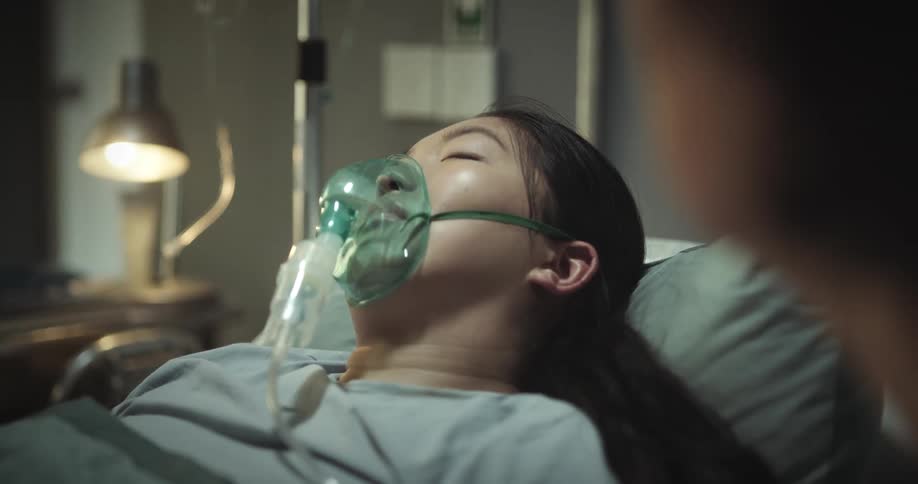 Mother holding and kissing the hands of her daughter who is sleeping with an oxygen mask in the hospital