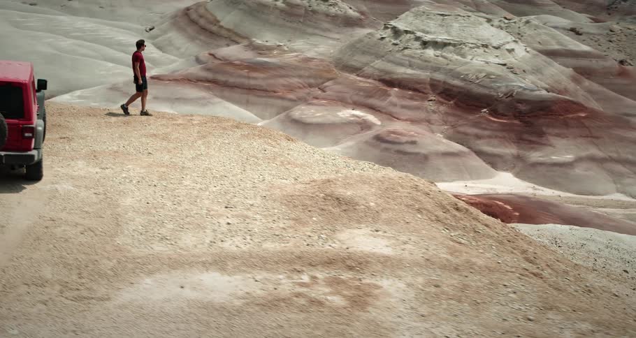 A man stands near the edge of a cliff in the desert on a sunny day with a red car parked behind him