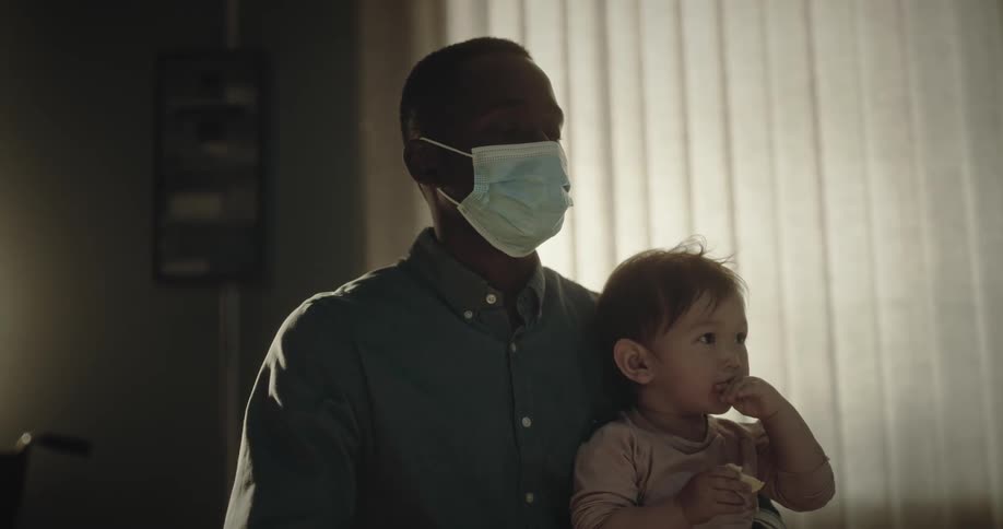 A male physician wearing a mask holds a young child in his arms