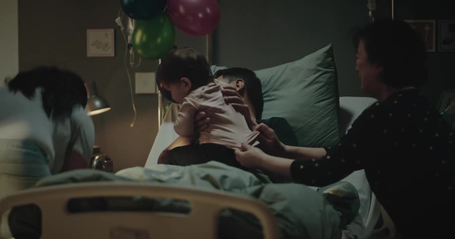 A man lying in a hospital bed playing with a child and his family around him