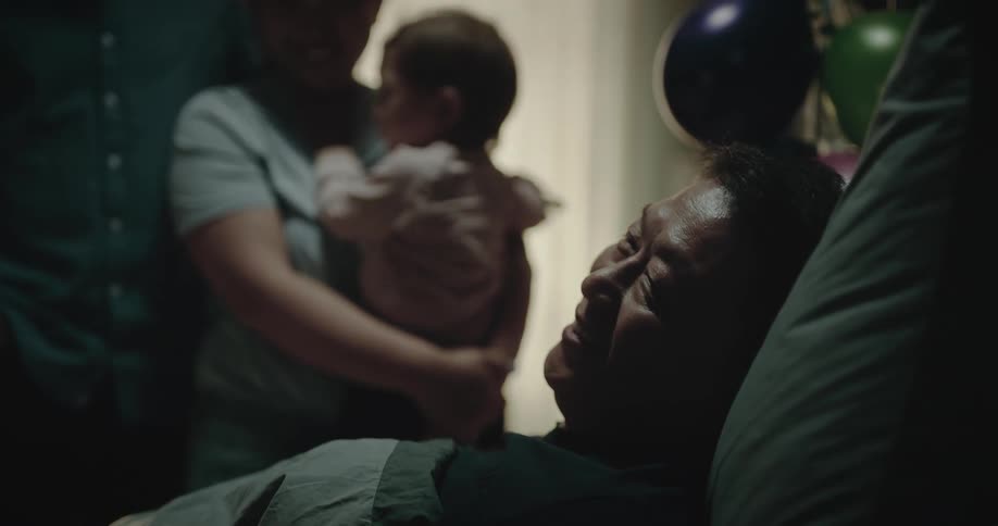 Woman holding baby stands near man lying in bed with balloons