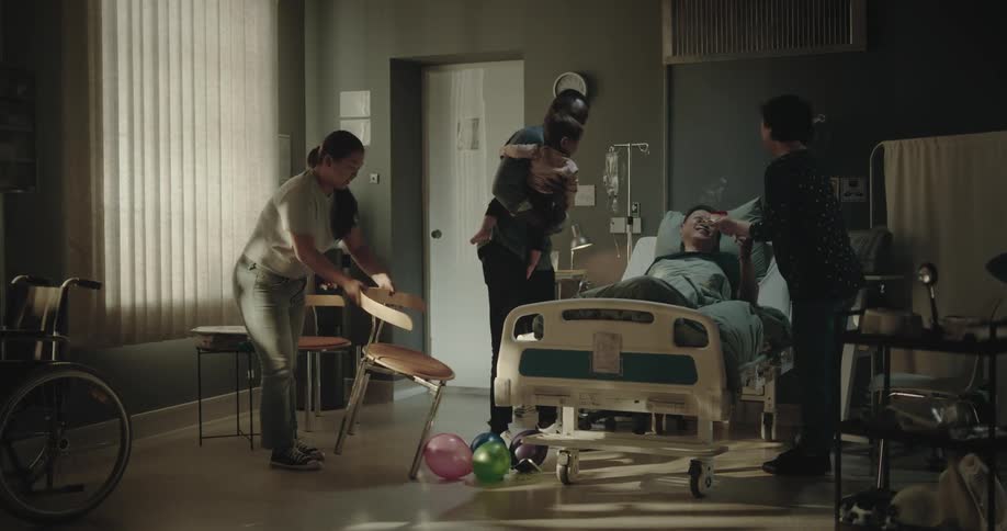Family coming into a room and singing happy birthday with a small cake for a person lying in the hospital bed