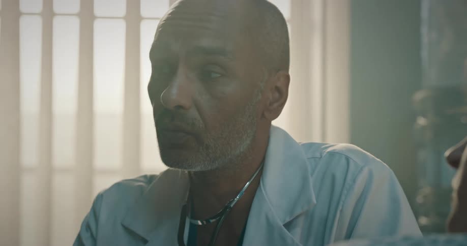 Male doctor wearing a lab coat talks to a patient lying in hospital bed and shakes his head