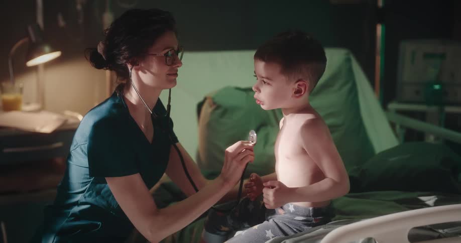 Nurse gives child checkup as boy sits on hospital bed at home