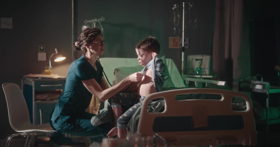 A doctor uses her stethoscope on a young boy sitting on a hospital bed