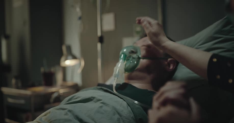 Woman holds hand of man lying in hospital bed with oxygen mask