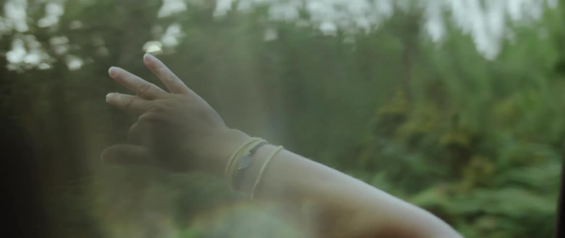 Woman moves her arm through air while traveling past woods during a road trip on a sunny day