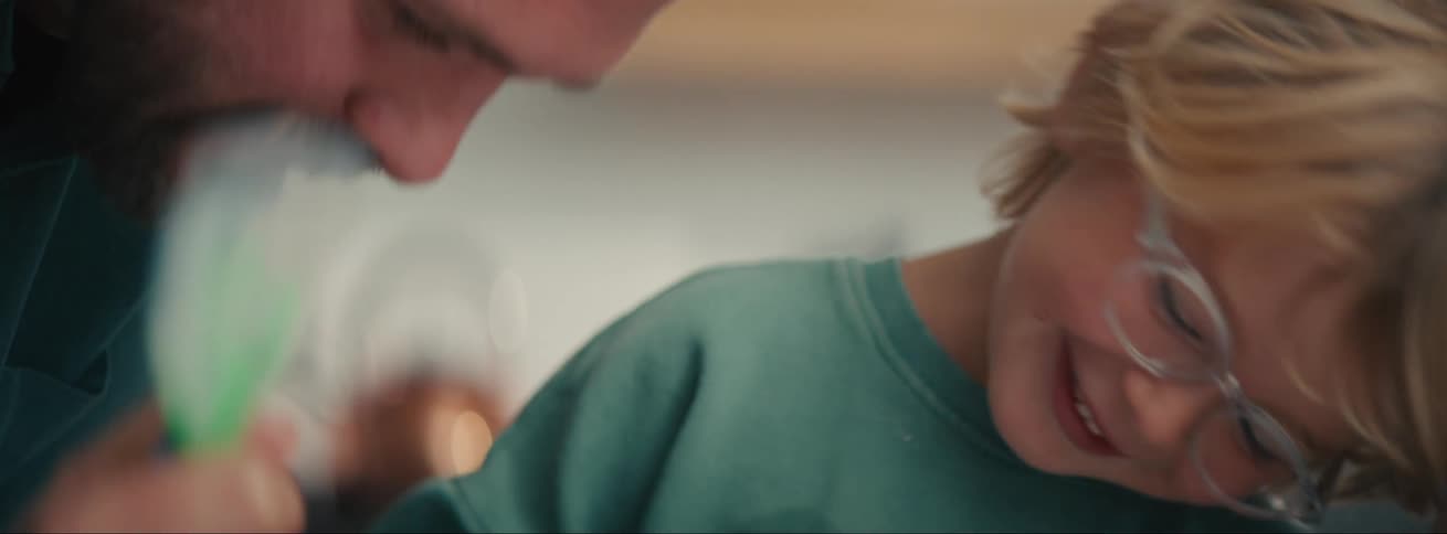 A little blond boy wearing glasses is smiling looking at something an adult is doing