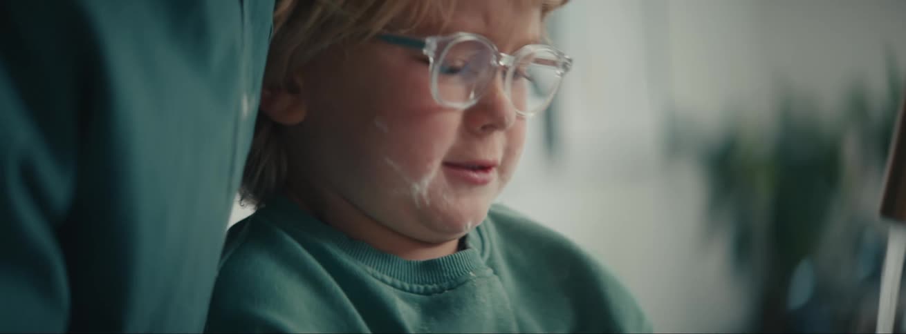 A little boy has an adult next to him washing his hand under a tap and there is some shaving cream on the boys face