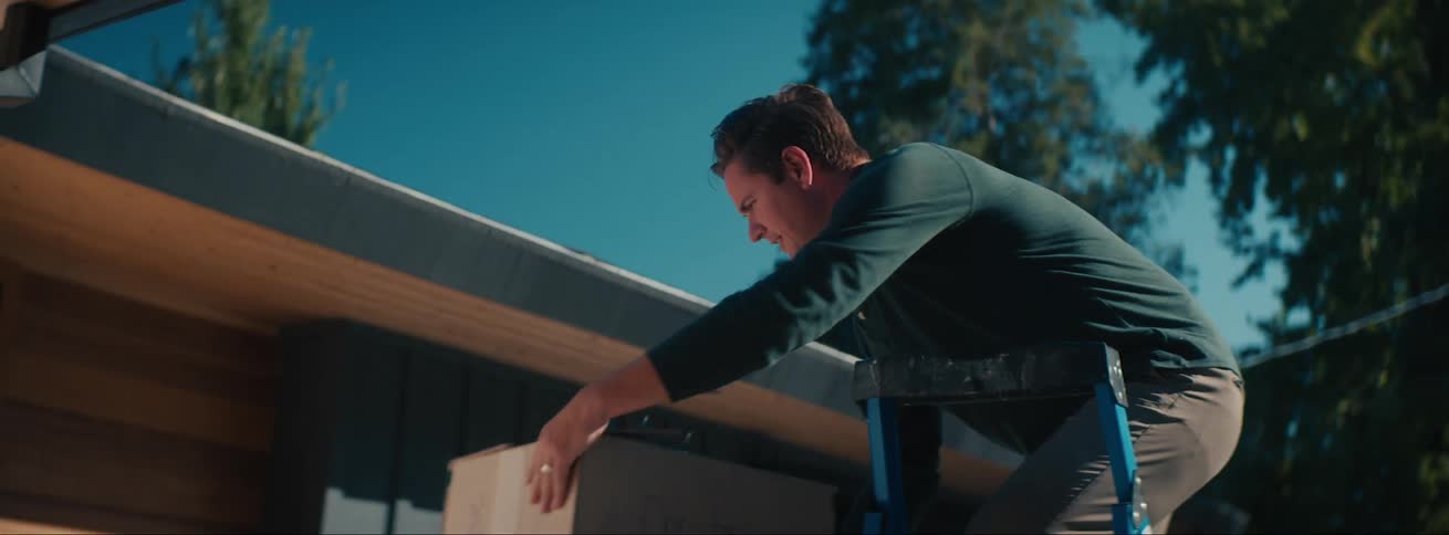 A father stands outside on a ladder while his son brings him a box of christmas lights