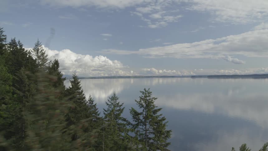 Flying through the trees of a coastal forest to reveal a glass calm bay reflecting the clouds and blue sky
