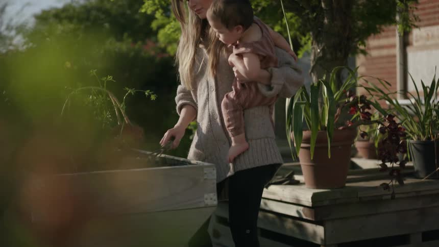 Mother holding her baby daughter while using a rake to tend to the soil in a small garden
