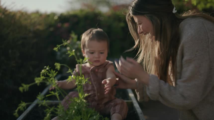 Woman places toddler in raised garden bed with plant