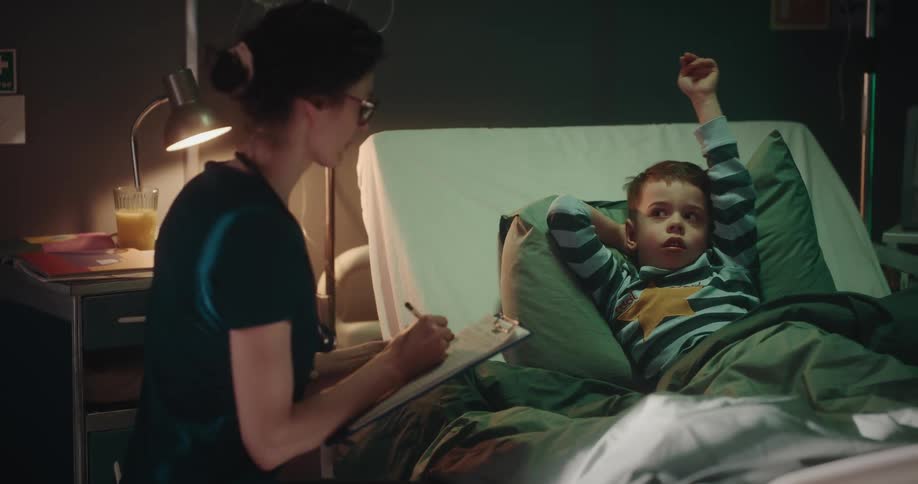 A nurse seated next to a boy in a hospital bed engaging in conversation and recording details in the medical records