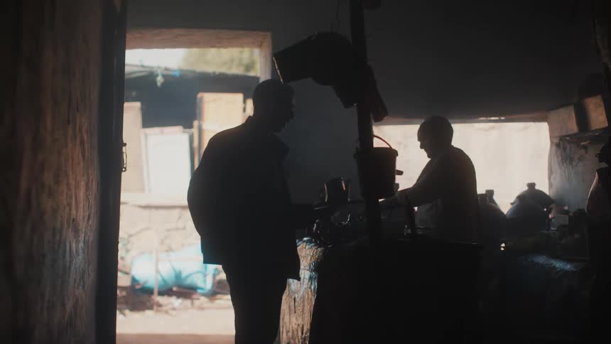 Man holding a kettle in a small shack with an older gentleman in front of him and a child in the background