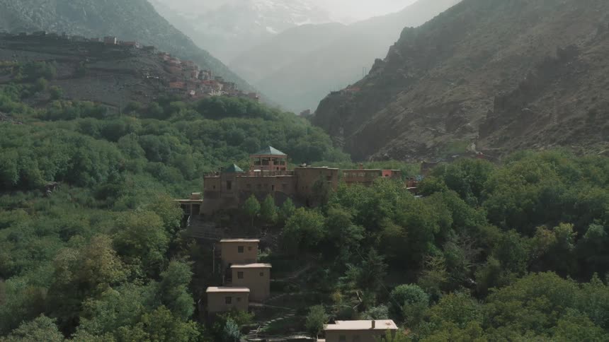 A building in the middle of a green forest and behind several mountains