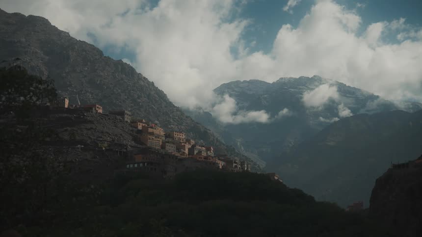 A village on a rocky mountain near two higher mountain peaks enveloped by clouds