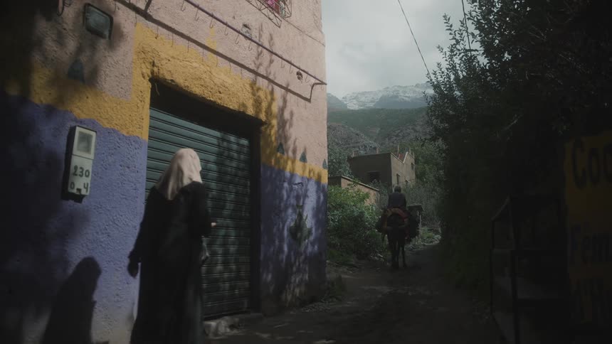 Rider on horseback and two female pedestrians pass building on path leading toward mountains