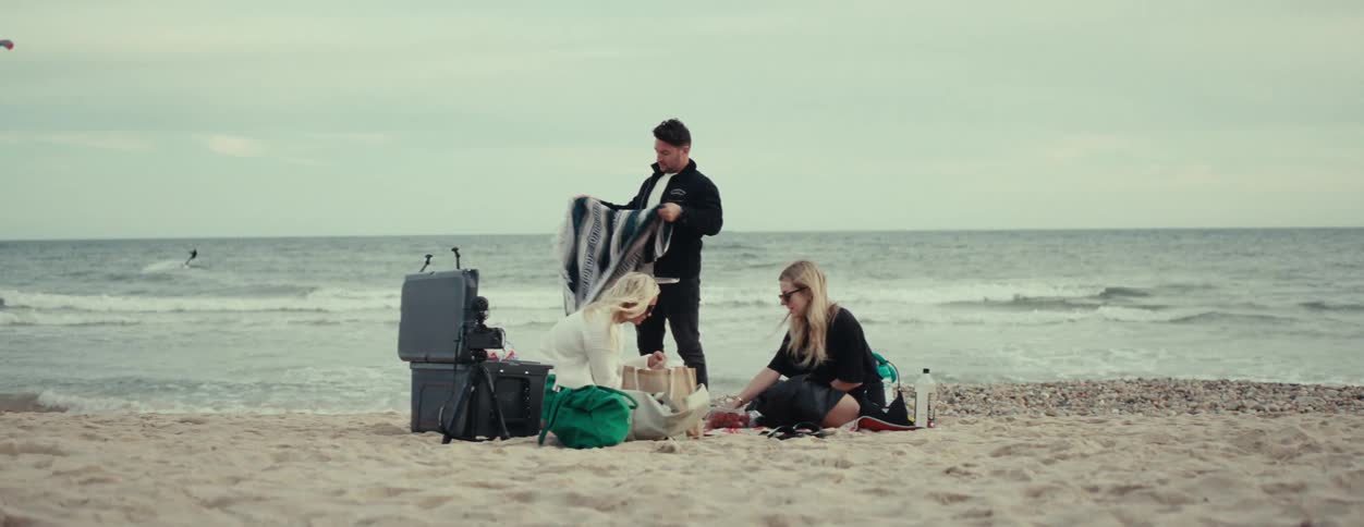 Three people are accommodated in front of a sea on the beach and are wearing warm clothes