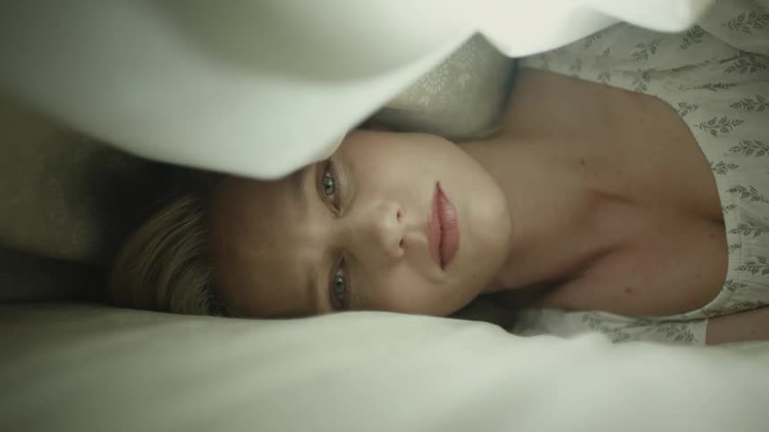 A beautiful woman wearing a nightgown is lying on her sidebetween 2 offwhite sheets she is looking straight ahead at the camera at first it is dark and then bright light spills over her