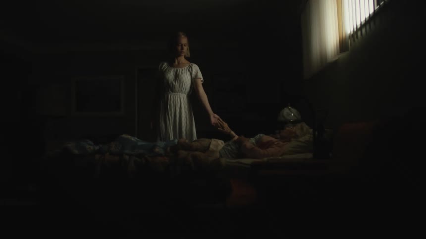 Woman in a white dress holding a elderly womans hand in a dark room
