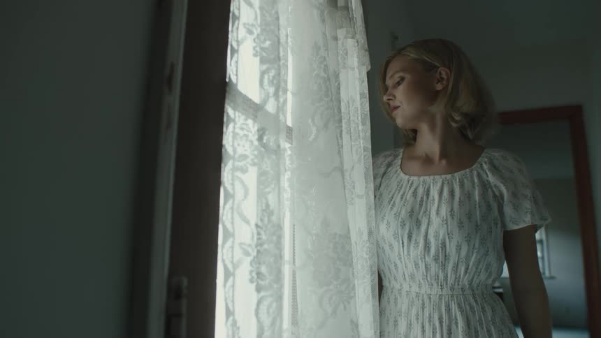 Woman stands looking out window through lacy curtain