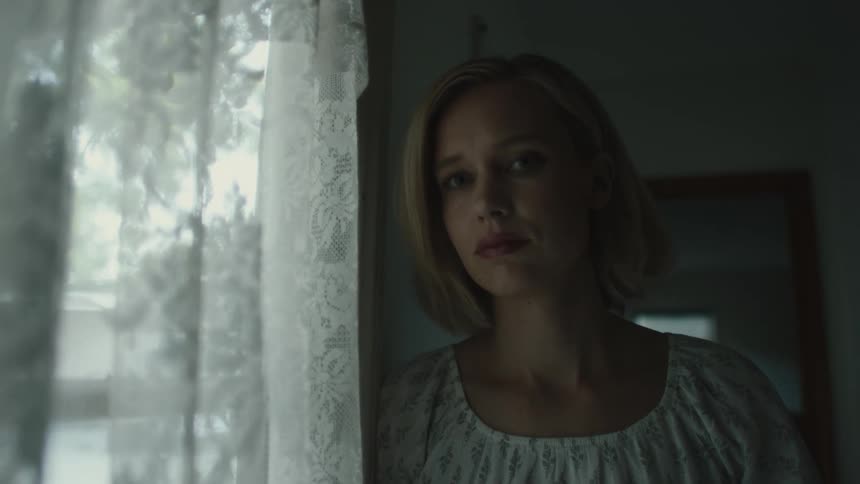 A woman with blonde hair wearing a white dress stands next to window curtains