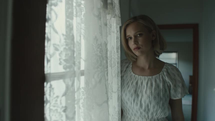 A woman with blonde hair dressed in white looks out the window with a somber expression
