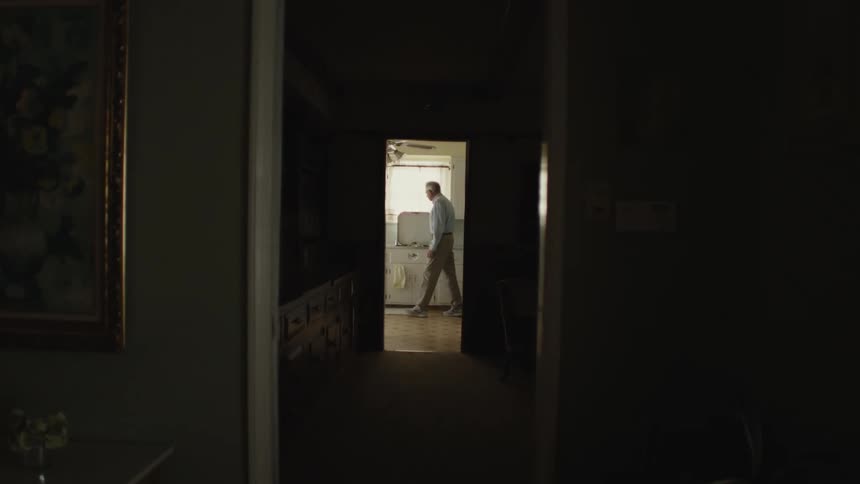 Elderly man leans against cabinet at end of series of rooms