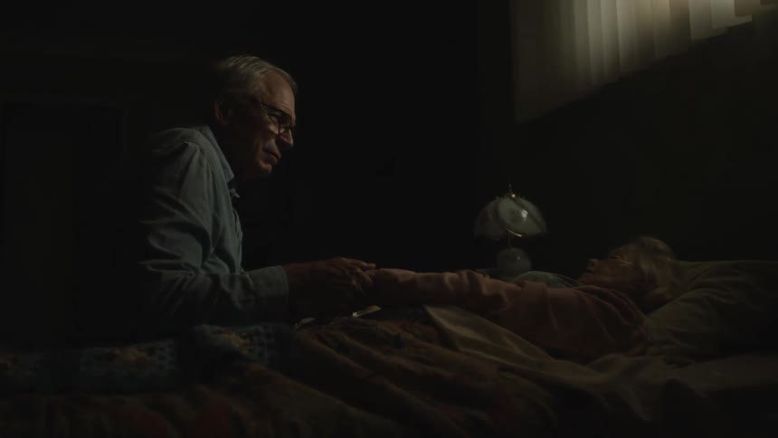 Elderly man sitting next to his wife lying in bed holding her hands