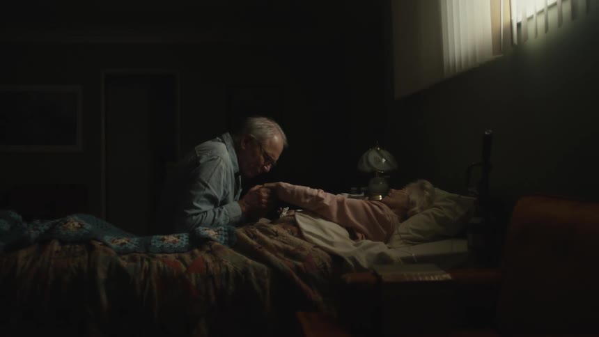 Light brightens then fades on elderly man kissing hand of elderly female invalid lying in bed