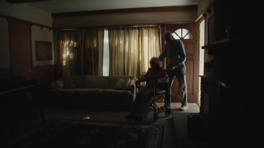 An elderly couple are entering through a door in a house and the woman is in a wheelchair