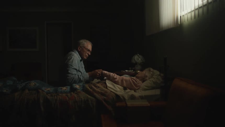 A man holding the hand of a woman who is lying down and both are elderly