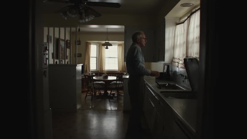 An elderly man wearing glasses a blue shirt and pants takes a glass of water from the kitchen sink