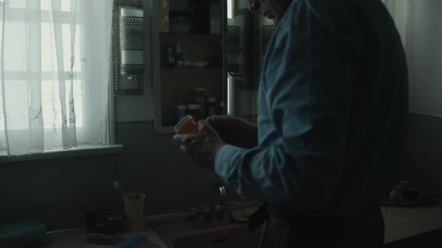 An elderly man with glasses wearing a blue shirt takes medicine in the bathroom