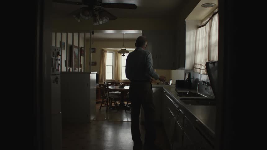 An old man walks to a tap and then stands in the kitchen and remains still
