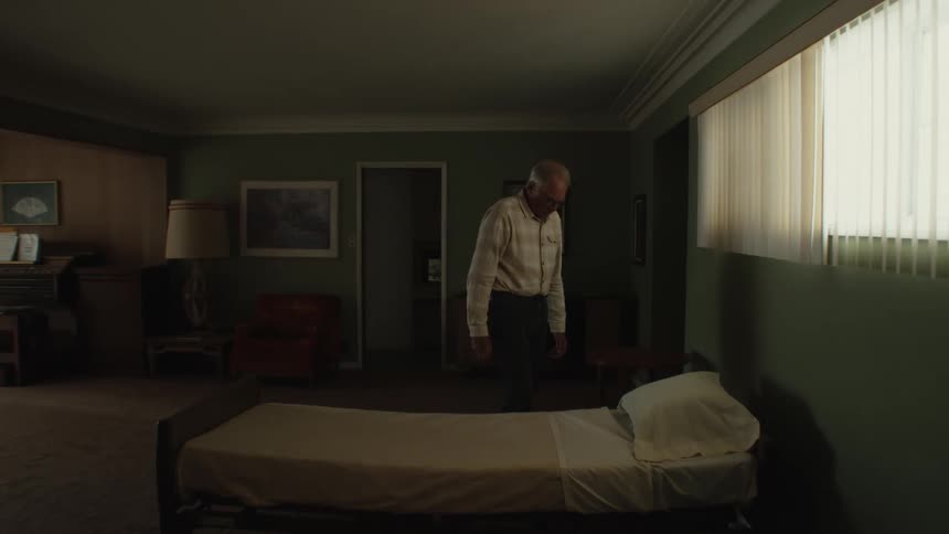Elderly man with glasses a white shirt and black pants walks around the bed in the room