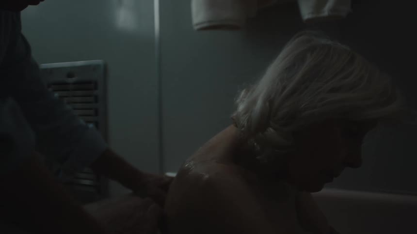 An elderly man rubs the back of an old woman as she sits in the bathtub